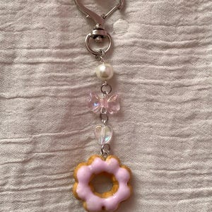 May include: A keychain with a silver heart-shaped clasp and a series of charms. The charms include a pearl, a pink bow, a heart-shaped bead, and a pink frosted donut charm. The donut charm has a light brown base and pink frosting.
