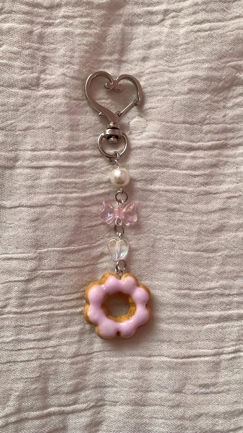 May include: A keychain with a silver heart-shaped clasp and a series of charms. The charms include a pearl, a pink bow, a heart-shaped bead, and a pink frosted donut charm. The donut charm has a light brown base and pink frosting.