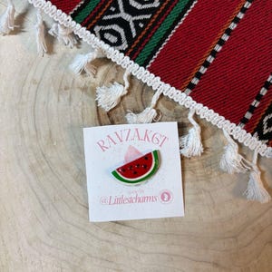 Può includere: Una spilla a forma di anguria con un design rosso, verde e nero e un cappello rosa. La spilla è su una carta bianca con il testo "RAVZAK6T" e "@Littlestchams". Lo sfondo include un tessuto a motivi rossi, verdi e neri.
