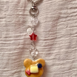 May include: A silver-colored keychain with a heart-shaped clasp. The keychain features clear beads, a red star, and a charm shaped like a pancake with butter, a blueberry, and a strawberry.