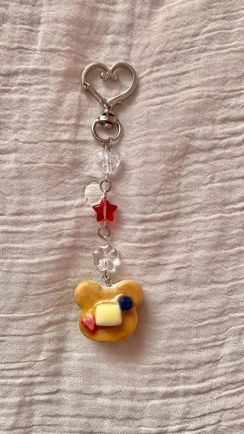 May include: A silver-colored keychain with a heart-shaped clasp. The keychain features clear beads, a red star, and a charm shaped like a pancake with butter, a blueberry, and a strawberry.