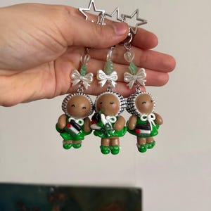 Op de afbeelding: Drie handgemaakte poppenoorbellen met een stervormige bovenkant. Elke pop heeft een bruin gezicht, een wit en zwart geruite hoed, een groene jurk en een witte strik. De oorbellen worden door een hand vastgehouden.