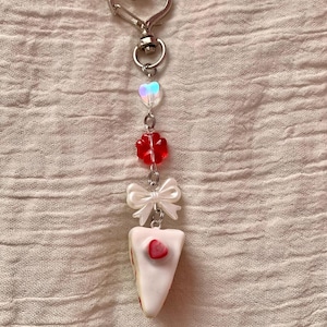 May include: A silver-toned keychain with a heart-shaped clasp. The charm features a series of beads, including a heart, a red flower, a white bow, and a slice of cake with a strawberry on top. The background is a textured, light pink fabric.