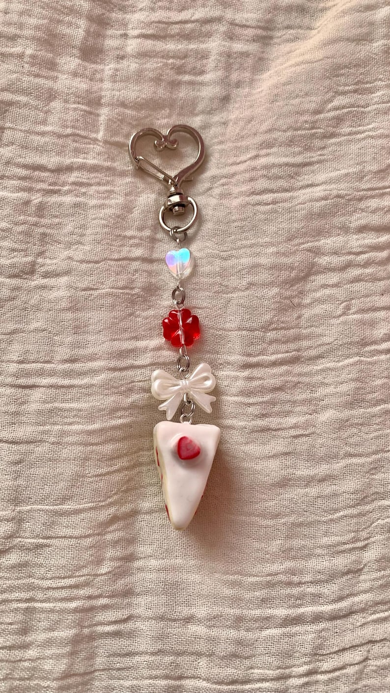 May include: A silver-toned keychain with a heart-shaped clasp. The charm features a series of beads, including a heart, a red flower, a white bow, and a slice of cake with a strawberry on top. The background is a textured, light pink fabric.