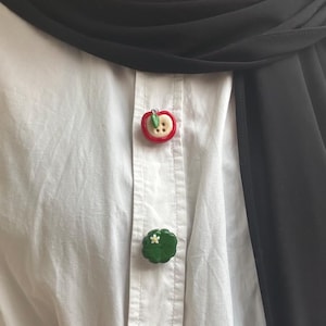 Op de afbeelding: Een wit overhemd met twee decoratieve knopen. De bovenste knoop is een rode appel met een groen blad en een beige knoop. De onderste knoop is een groene, geschulpte vorm met een kleine witte bloem.