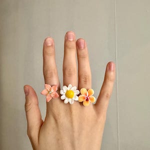 Könnte beinhalten: Drei blütenförmige Ringe in Pfirsich-, Weiß- und Orangetönen werden auf einer Hand gezeigt. Der Pfirsichring hat einen kleinen Kristallakzent. Der weiße Ring hat eine gelbe Mitte. Der orangefarbene Ring hat rote Akzente.