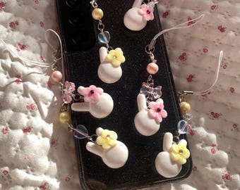 Puede incluir: Una funda de teléfono negra con múltiples colgantes de conejito blancos, cada uno adornado con una flor rosa o amarilla. Los colgantes están conectados por cadenas plateadas y cuentas coloridas. La funda del teléfono descansa sobre una superficie con estampado floral.