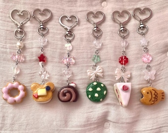 Puede incluir: Seis llaveros con cierres plateados en forma de corazón. Cada llavero presenta un dije de comida en miniatura diferente, que incluye una rosquilla, un panqueque con forma de oso, un brazo de gitano, una galleta verde con lunares, una rebanada de pastel y un pescado.