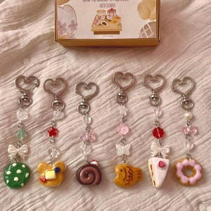 Peut inclure: Ensemble de six porte-clés avec des miniatures de pâtisseries japonaises, dont un mochi vert, une crêpe en forme d'ours et un gâteau roulé. Chaque porte-clés est orné d'un fermoir en forme de cœur, de perles et de breloques décoratives. Une boîte "Blind Box Japanese Pastry Keychains" est également présente.