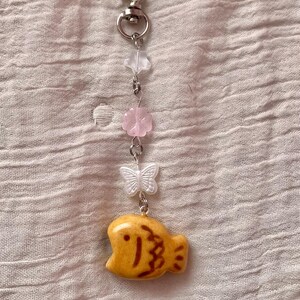 May include: A keychain with a heart-shaped clasp, featuring a series of charms. The charms include a clear star, pink flower, white butterfly, and a yellow fish-shaped charm with brown details. The keychain is made of metal.