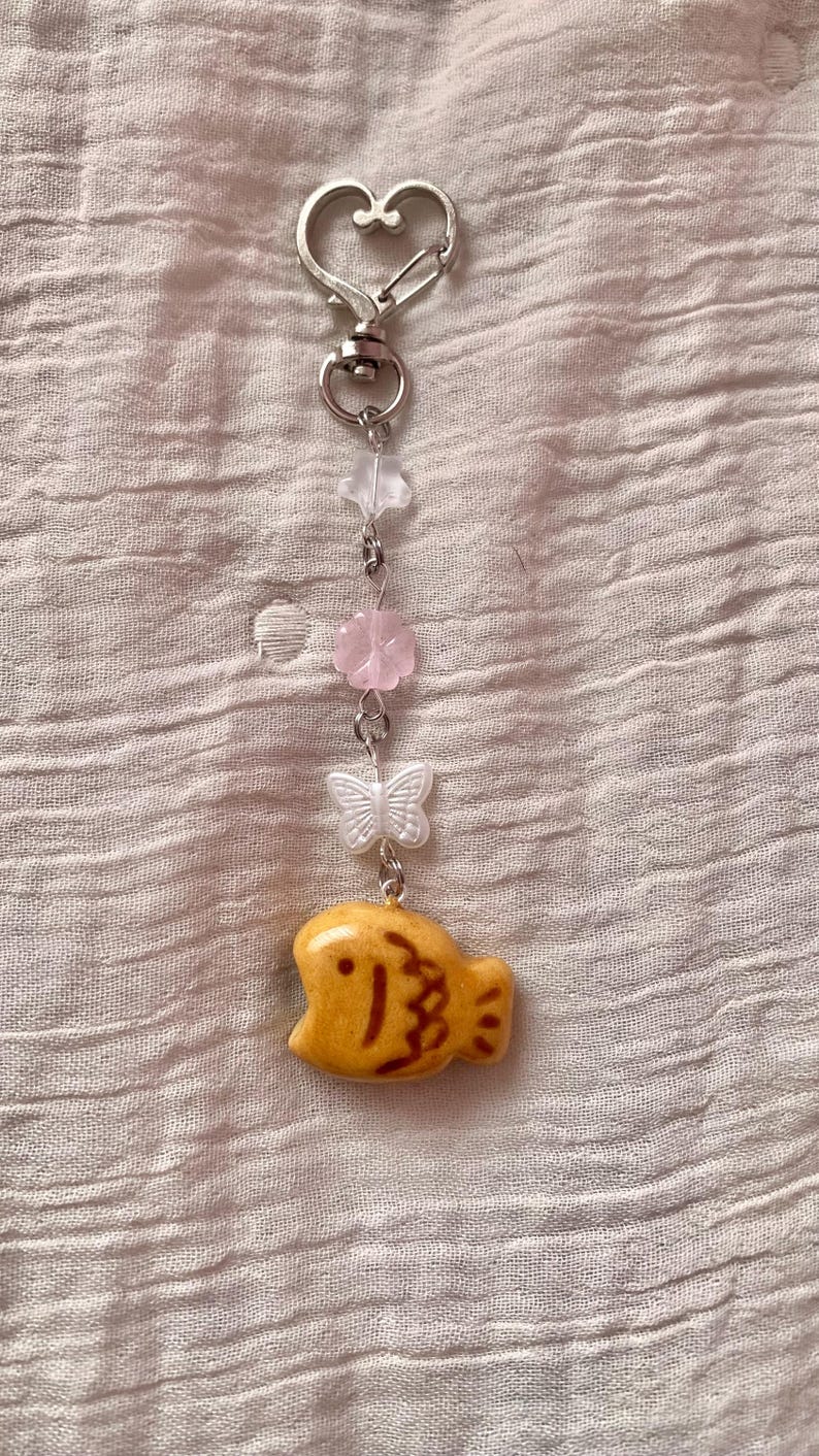 May include: A keychain with a heart-shaped clasp, featuring a series of charms. The charms include a clear star, pink flower, white butterfly, and a yellow fish-shaped charm with brown details. The keychain is made of metal.