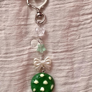 May include: A handmade keychain with a silver heart-shaped clasp. The keychain features clear heart-shaped beads, a green star-shaped bead, a white bow, and a green, polka-dotted charm.