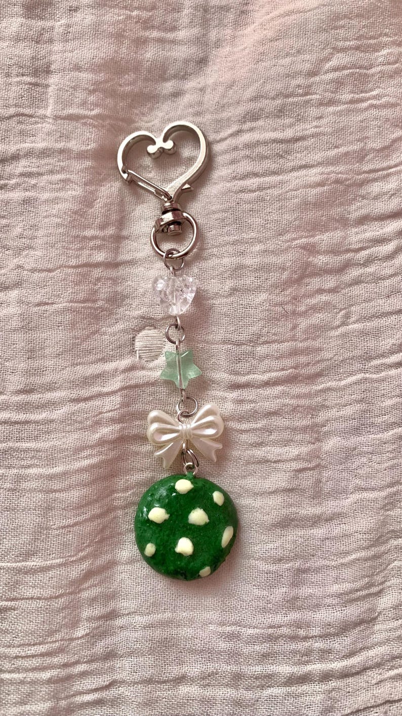May include: A handmade keychain with a silver heart-shaped clasp. The keychain features clear heart-shaped beads, a green star-shaped bead, a white bow, and a green, polka-dotted charm.