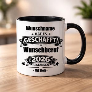 Könnte beinhalten: Weißer Keramikbecher mit schwarzem Innenraum und Henkel. Der Becher trägt den schwarzen Text "Wunschname HAT ES GESCHAFFT! Wunschberuf 2026 BESTANDEN - Mit Stolz". Der Becher steht auf einer Holzoberfläche.