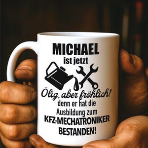 Könnte beinhalten: Weißer Keramikbecher mit dem Text "MICHAEL ist jetzt Ölig, aber fröhlich! denn er hat die Ausbildung zum KFZ-MECHATRONIKER BESTANDEN!" und einer Grafik von Schraubenschlüssel und Ölkanne. Der Becher wird von Händen gehalten.
