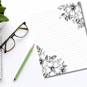 May include: A white notepad with horizontal lines and floral corner designs in black and white. A green pencil, black-framed glasses, and a small succulent plant in a white pot are also on the white surface.