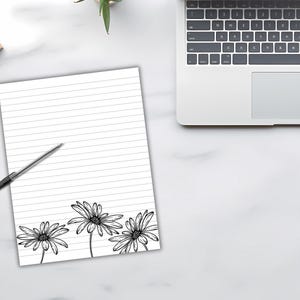 May include: A lined white notepad with a black pen and daisy floral design. The notepad is on a white surface next to a laptop. The daisy design is at the bottom of the notepad.