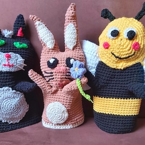 Könnte beinhalten: Drei gehäkelte Fingerpuppen: eine schwarze Katze, ein rosa Kaninchen, das eine lila Blume hält, und eine gelbschwarze Biene.