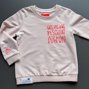 May include: A pale pink sweatshirt with long sleeves. The text "Lass uns was im Schilde führen!" is printed in red on the front. A red smiley face is on the left sleeve. A small label is at the bottom.