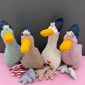 Könnte beinhalten: Vier Plüsch-Gänse mit unterschiedlichem Fell, die blaue Strickmützen tragen. Die Gänse sitzen auf einer rosa Oberfläche mit ausgestreckten Füßen. Jede Gans hat ein Herz in einer anderen Farbe auf der Brust.