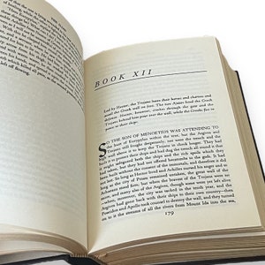 May include: An open book with a black cover, showing text from "The Iliad." The page is open to "Book XII," with black text. The page number "179" is visible at the bottom. The book is on a white surface.