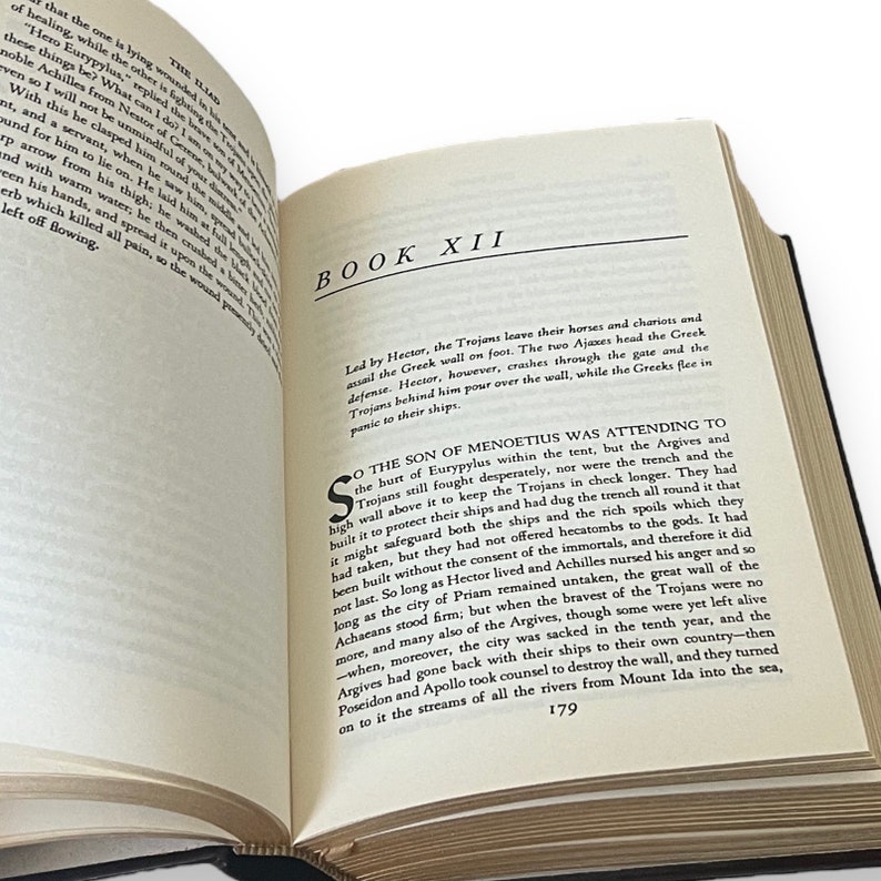 May include: An open book with a black cover, showing text from "The Iliad." The page is open to "Book XII," with black text. The page number "179" is visible at the bottom. The book is on a white surface.