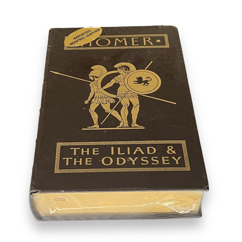 May include: A dark brown hardcover book, "The Iliad & The Odyssey" by Homer, with gold lettering and illustrations of two figures with spears and shields. The book is wrapped in clear plastic.