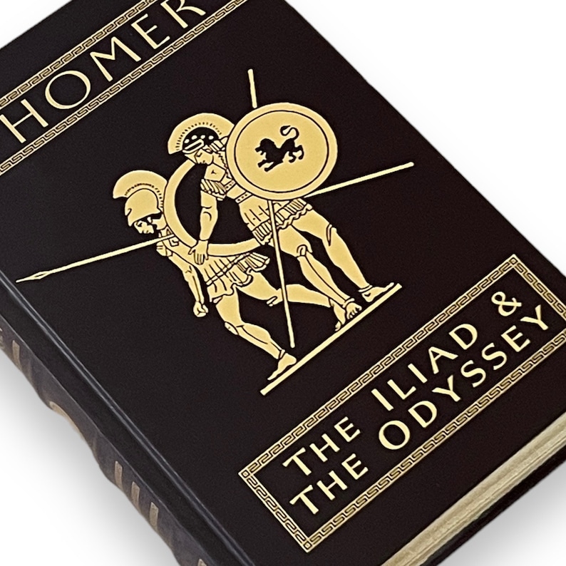 May include: A dark brown hardcover book featuring the title "The Iliad & The Odyssey" by Homer. Gold lettering and a gold illustration of Greek warriors adorn the cover. The book's spine is visible, and the pages are off-white.