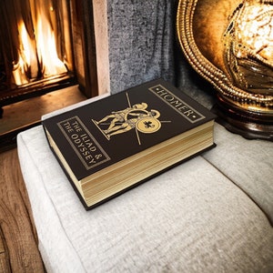 May include: A hardback book titled "The Iliad & The Odyssey" by Homer. The book has a dark brown cover with gold lettering and illustrations of warriors. The pages have a gold gilt edge. The book is resting on a light grey cushion.
