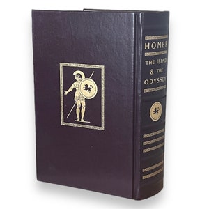 May include: A dark purple hardcover book titled "Homer The Iliad & The Odyssey". The spine and front cover have gold lettering and a gold illustration of a warrior with a shield. A classic literature edition.