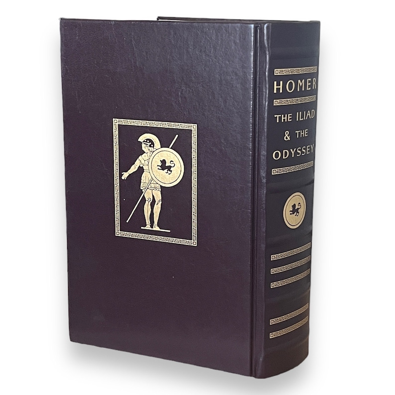 May include: A dark purple hardcover book titled "Homer The Iliad & The Odyssey". The spine and front cover have gold lettering and a gold illustration of a warrior with a shield. A classic literature edition.