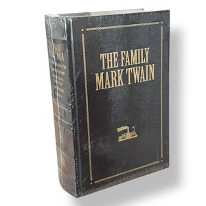 Op de afbeelding: Een zwart hardcover boek met de titel "The Family Mark Twain" met gouden letters en een stoomboot illustratie. Het boek is verpakt in doorzichtig plastic.