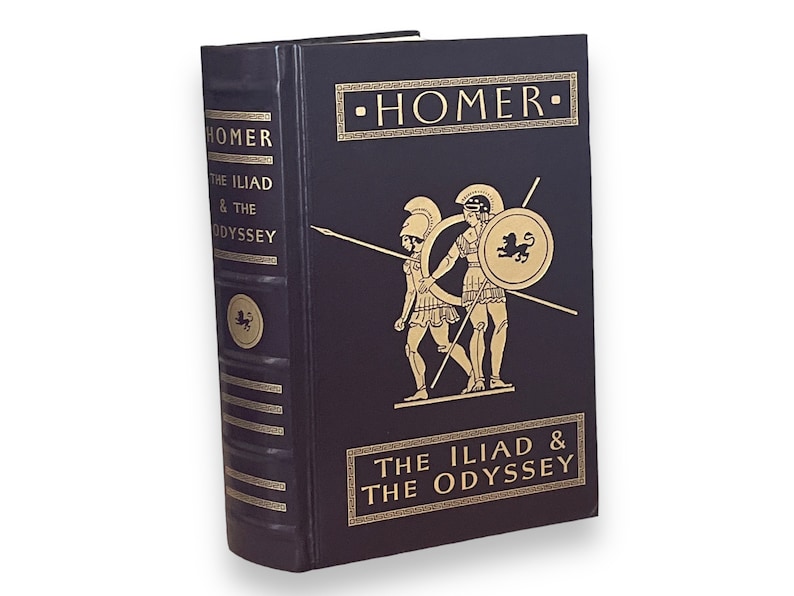 May include: A hardback book titled "Homer The Iliad & The Odyssey." The dark blue book features gold lettering and illustrations of Greek warriors. The spine and cover have decorative gold accents, and a gold emblem.
