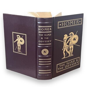 May include: A burgundy hardcover book titled "Homer The Iliad & The Odyssey". Gold lettering and illustrations of Greek figures are on the cover and spine. The book is open, revealing the pages.