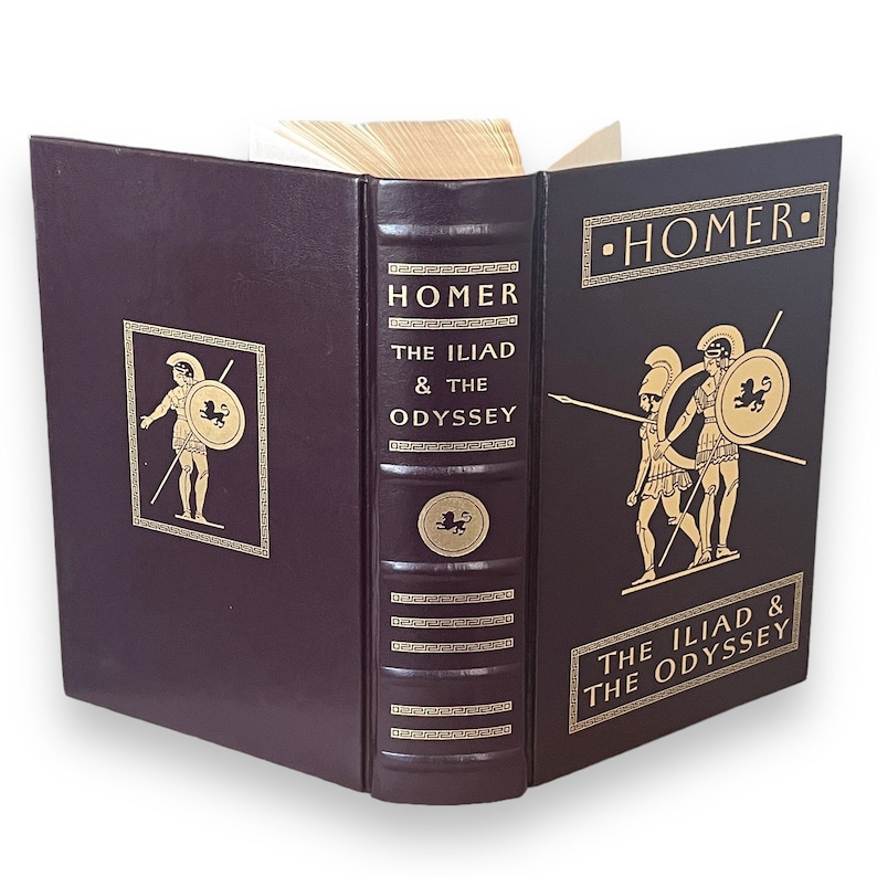 May include: A burgundy hardcover book titled "Homer The Iliad & The Odyssey". Gold lettering and illustrations of Greek figures are on the cover and spine. The book is open, revealing the pages.