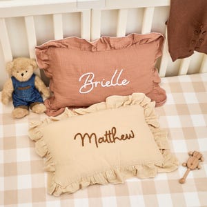 Può includere: Due cuscini rettangolari in mussola con bordi a volant. Il cuscino superiore è rosa polvere con il nome Brielle ricamato in corsivo bianco. Il cuscino inferiore è beige con il nome Matthew ricamato in corsivo marrone scuro. Un orsacchiotto è posizionato accanto.