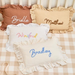 May include: Four rectangular muslin pillows with ruffled edges, each featuring embroidered names. The top left is terracotta with white text 'Brielle', top right is beige with brown text 'Matthew', bottom left is white with multicoloured text 'Winifred', and bottom right is cream with blue text 'Bradley'.