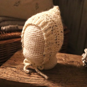 May include: A white crocheted baby bonnet with a lace trim. The bonnet is tied with a white ribbon and is displayed on a wooden surface.