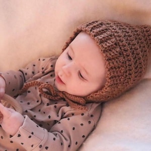 May include: A baby wearing a brown knitted hat with a pointed tip and a brown and white polka dot onesie. The baby is holding a brown and white stuffed animal.