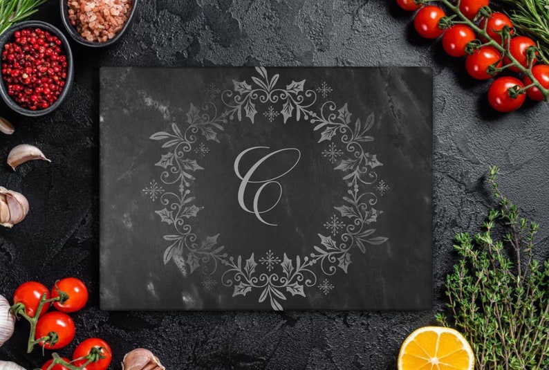 May include: A black slate serving board with a decorative engraved design of a holly wreath and snowflakes. The wreath has a letter "C" in the center. The board is surrounded by various food items, including red peppercorns, tomatoes, garlic, and herbs.