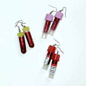 May include: Three pairs of drop earrings featuring miniature clear plastic laboratory tubes filled with red glitter. The tubes have yellow, lavender, and red screw-on caps. Each earring is attached to a silver-toned metal fishhook ear wire.