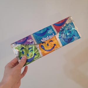 May include: A colorful zippered pouch made from recycled Kool-Aid packets. The pouch features a variety of flavors including Tropical Punch, Grape, Strawberry Kiwi, and Blue Raspberry.