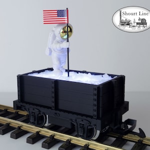May include: A black toy train wagon with a white astronaut figurine holding an American flag. The wagon is filled with white, crushed ice-like material. The wagon is on a set of train tracks.