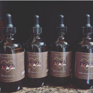 May include: Four brown glass bottles of hair growth oil with black droppers. Each bottle has a brown label with a heart-shaped graphic of women and the text "Hair Growth Oil" and "Scalp Massage Oil". The brand name "The Urban Hippie Patch" is also visible.