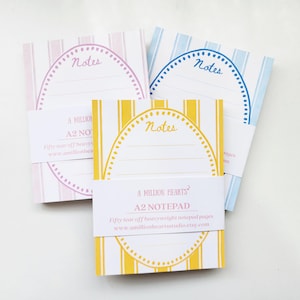 Peut inclure: Trois blocs-notes A2 aux couleurs pastel : rose, bleu et jaune. Chaque bloc-notes présente un motif à rayures et le mot "Notes" en écriture manuscrite. Les blocs-notes sont maintenus ensemble par une bande de papier avec "A MILLION HEARTS" et "A2 NOTEPAD".