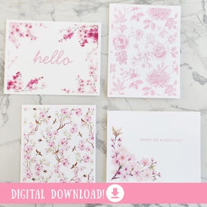 May include: Four white greeting cards with floral designs. One card says "hello" in pink script, framed by pink cherry blossoms. Another features a pink floral pattern. The third has a cherry blossom branch design, and the fourth says "SORRY WE MISSED YOU!"