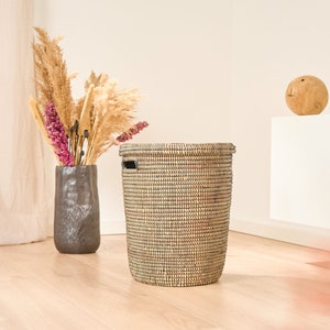 May include: A woven black and tan storage basket with a black handle. The basket is sitting on a light wood floor.