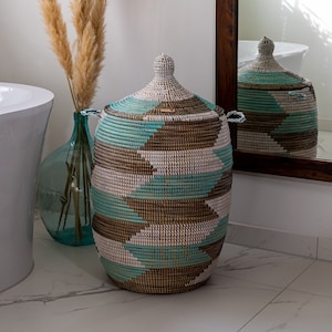 Puede incluir: Una cesta de almacenamiento grande y tejida con tapa. La cesta est&aacute; hecha de materiales naturales y tiene un patr&oacute;n geom&eacute;trico en tonos marrones, blancos y azul verdoso.