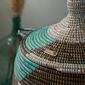 Puede incluir: Una cesta grande, redonda y tejida con tapa. La cesta est&aacute; hecha de materiales naturales y tiene un esquema de color negro, blanco y turquesa. La cesta tiene una etiqueta de madera con la palabra "YAADU" en ella.