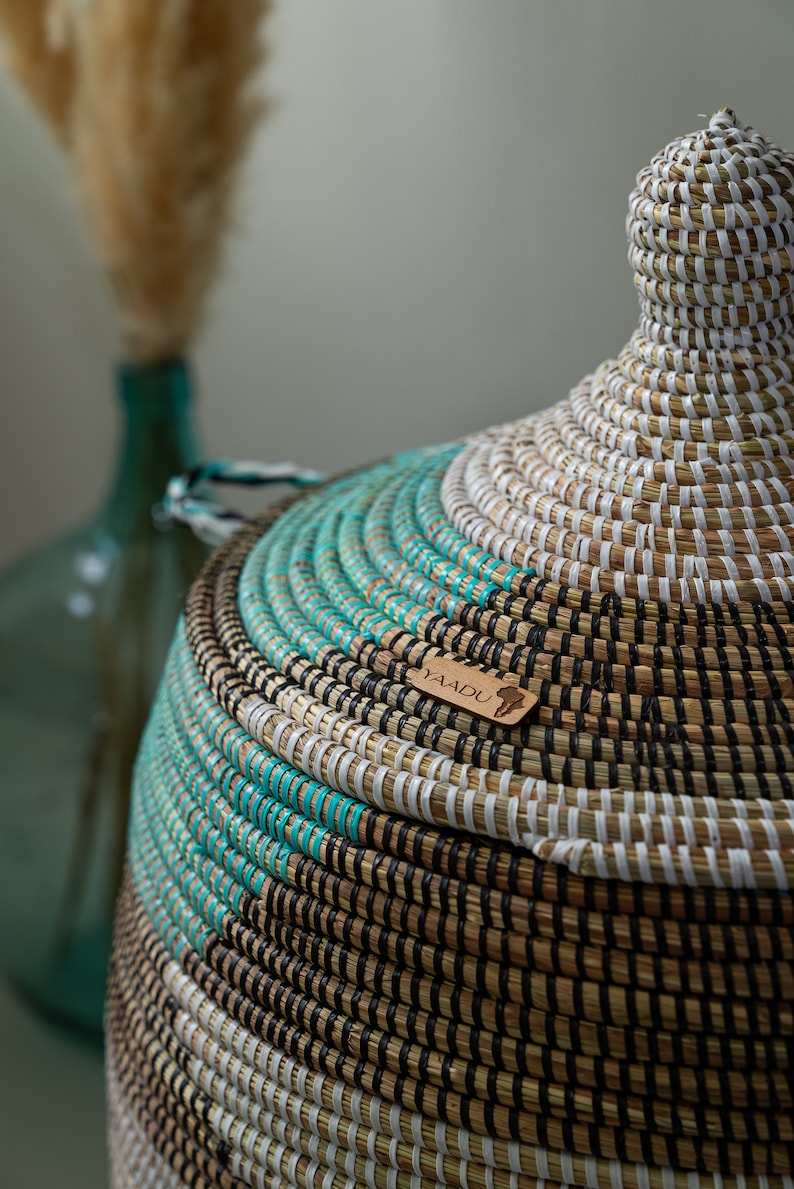 Puede incluir: Una cesta grande, redonda y tejida con tapa. La cesta est&aacute; hecha de materiales naturales y tiene un esquema de color negro, blanco y turquesa. La cesta tiene una etiqueta de madera con la palabra "YAADU" en ella.